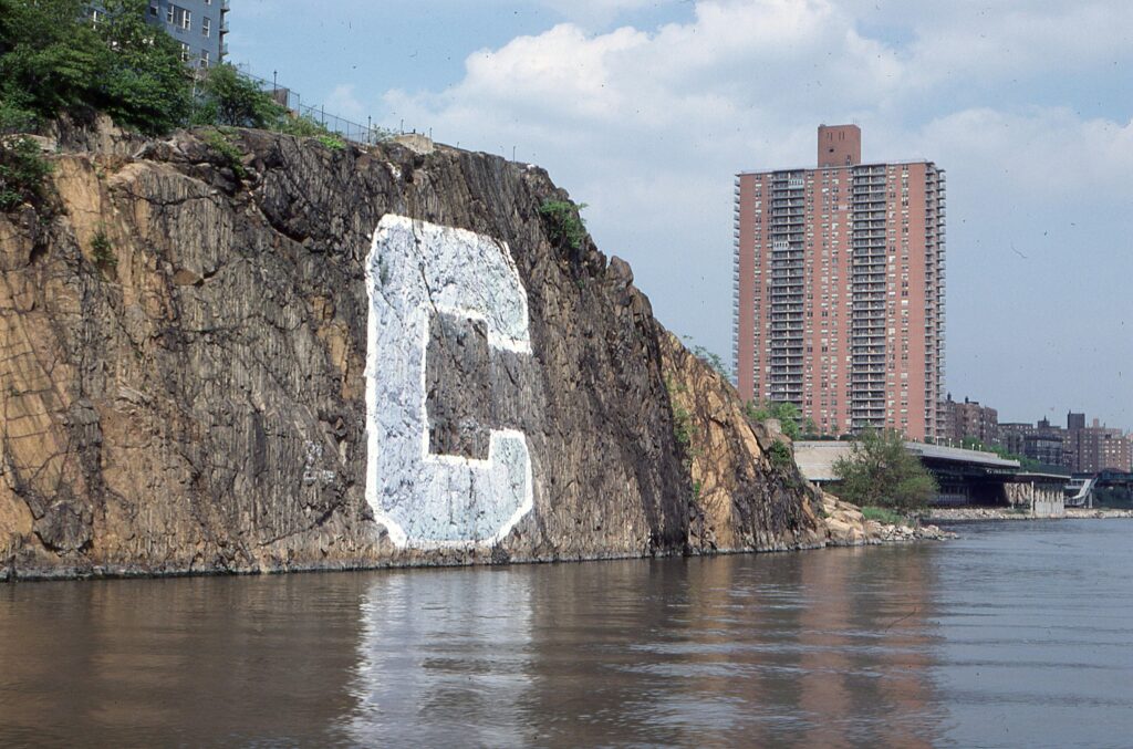 A letter 'C' painted on a riverside cliff next to urban high-rise buildings.