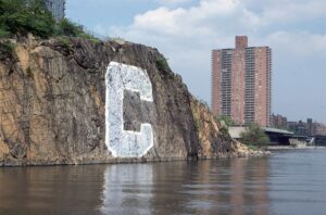 A letter 'C' painted on a riverside cliff next to urban high-rise buildings.