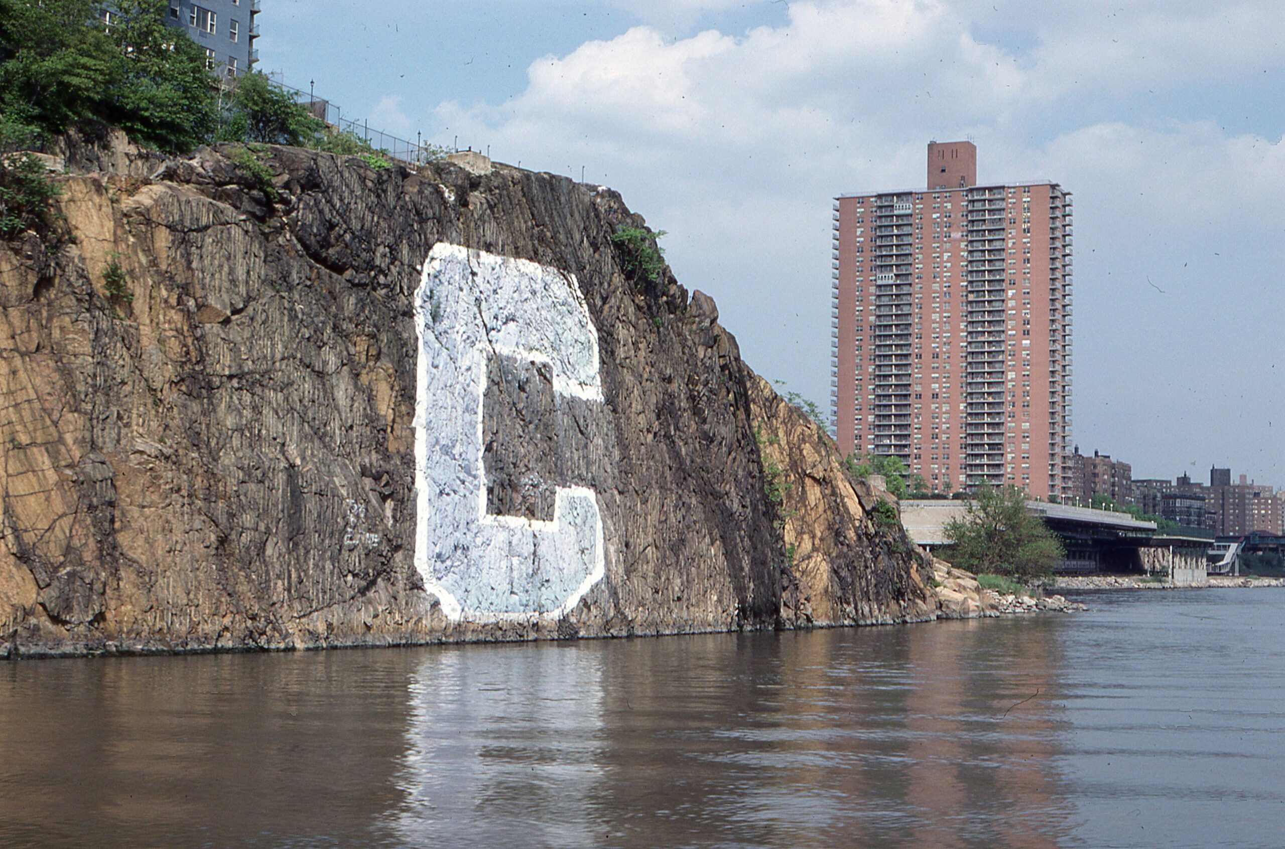 A letter 'C' painted on a riverside cliff next to urban high-rise buildings.