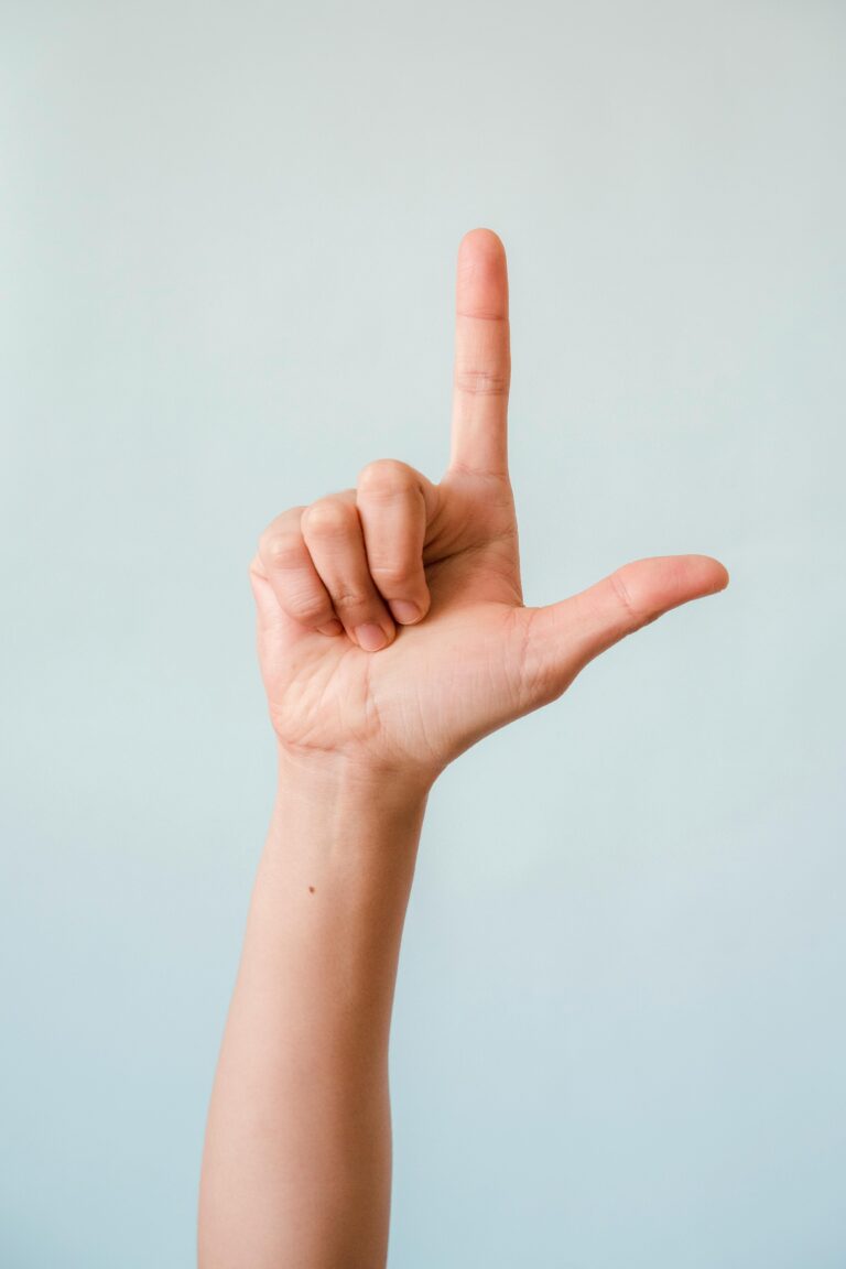 A close-up photo of a hand in an 'L' sign language gesture against a light background.